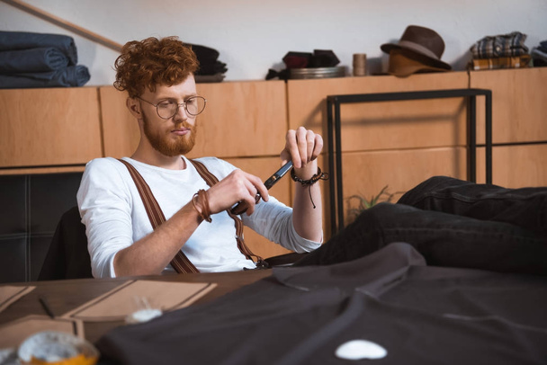 jonge mannelijke modeontwerper schaar te houden terwijl zittend aan tafel met naaiende patronen en stof - Foto, afbeelding