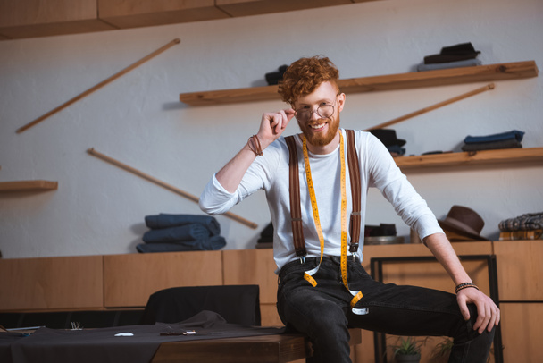 joven diseñador de moda masculino en gafas con cinta métrica alrededor del cuello sonriendo a la cámara en el lugar de trabajo
 - Foto, Imagen
