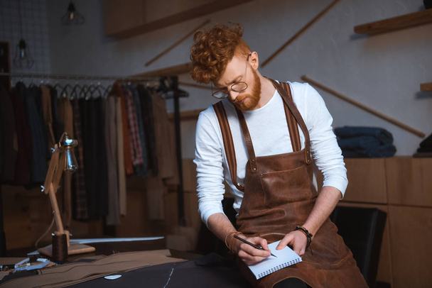 young male fashion designer in apron and eyeglasses taking notes in notebook  - Foto, afbeelding