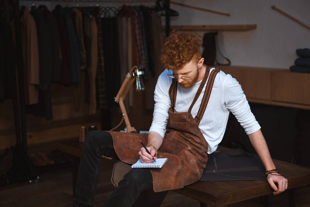 young male fashion designer in apron and eyeglasses taking notes in notebook - Foto, Imagem