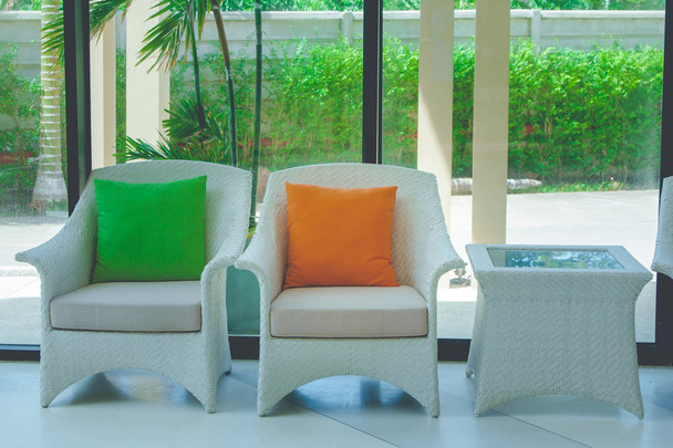 Groene en oranje hoofdkussens op wit weven stoel instelling op betonnen vloer in de lobby van hotel met groene bush buiten op de achtergrond. - Foto, afbeelding