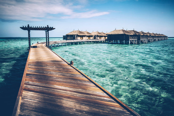 Jetty de madeira com arco em uma água do oceano azul-turquesa limpa. Moradias aquáticas. Viagens de verão em Maldivas
 - Foto, Imagem