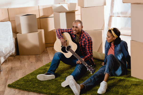 african american man playing on acoustic guitar for girlfriend in new apartment with cardboard boxes - Photo, Image