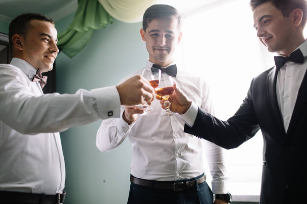 groom and his groomsmen friends in stylish suits drinking whiskey in hotel room, morning before the wedding preparation, emotional group of friends celebrating - Foto, immagini