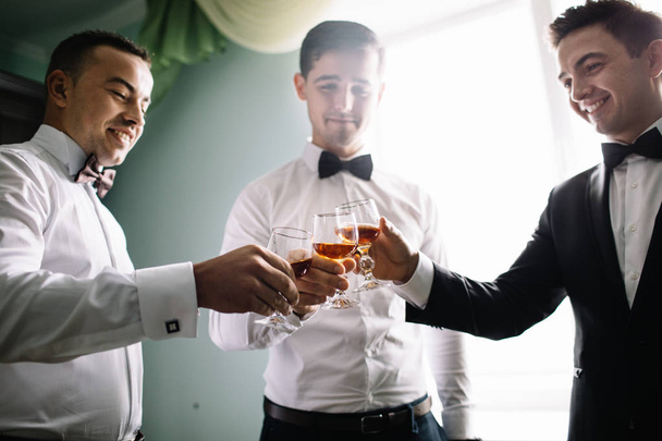 pana młodego i jego przyjaciele drużbami w stylowe kombinezony picie whisky w pokoju hotelowym, rano przed przygotowaniem ślubu, emocjonalne grupy przyjaciół z okazji - Zdjęcie, obraz