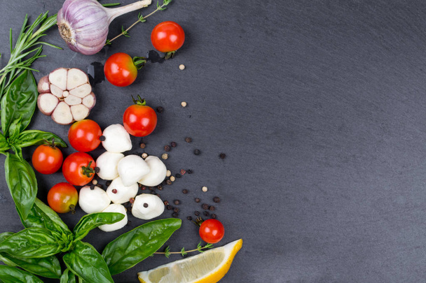 Mozzarella, cherry tomatoes and fresh basil, lemon, garlic. Ingredients for salad. On black slate stone chalkboard. Top view. - 写真・画像