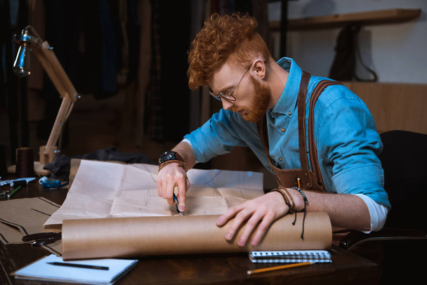 young male fashion designer in apron and eyeglasses making sewing patterns at workplace - Foto, afbeelding