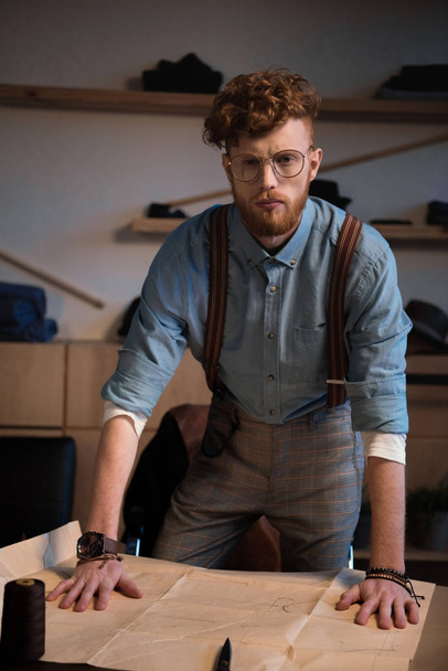 handsome young fashion designer in eyeglasses looking at camera while leaning at table with craft paper - Fotografie, Obrázek