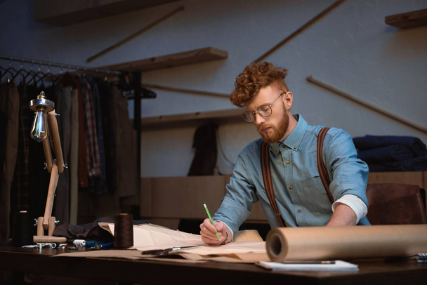 όμορφος νεαρά σχεδιαστής μόδας με γυαλιά εργασίας με σκάφη χαρτί στο εργαστήρι - Φωτογραφία, εικόνα