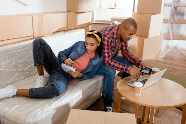 jovem Africano americano casal planejamento com bloco de notas e gadgets no sofá em nova casa
 - Foto, Imagem