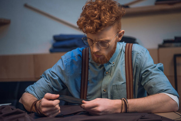 νεαρά αρσενικά σχεδιαστής μόδας στα γυαλιά εργασίας με ύφασμα στο τραπέζι - Φωτογραφία, εικόνα