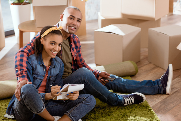 sorridente coppia afroamericana con smartphone e blocco note seduto nel nuovo appartamento
 - Foto, immagini