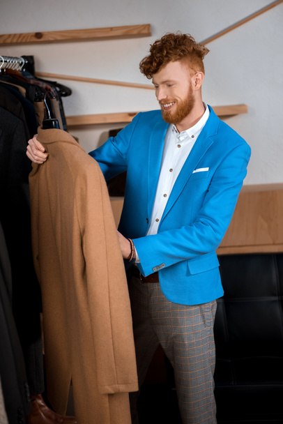 handsome smiling young man holding hanger with stylish brown overcoat in boutique - Φωτογραφία, εικόνα