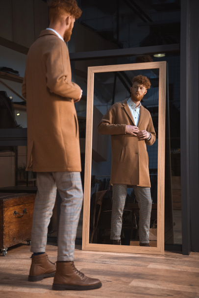 κομψό νεαρός άνδρας φοράει παλτό και κοιτάζοντας καθρέφτη στην μπουτίκ - Φωτογραφία, εικόνα