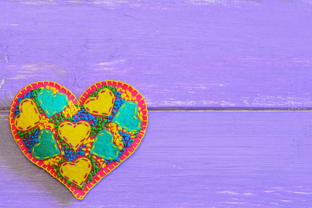 Decorative felt heart for Valentines Day background. Valentines Day hand embroidery pattern. Embroidered heart ornament DIY isolated on purple wooden background with copy space for text. DIY applique motif. Closeup - Photo, image
