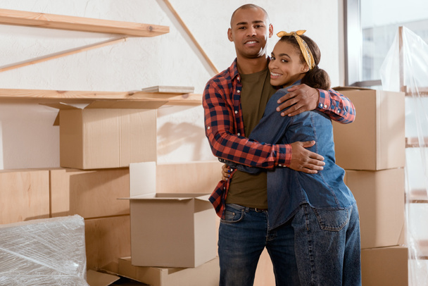 feliz casal afro-americano abraçando em novo apartamento com caixas de papelão
 - Foto, Imagem