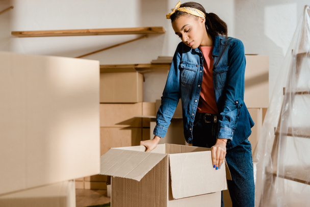 upset african american girl unpacking cardboard boxes in new apartment - Foto, imagen