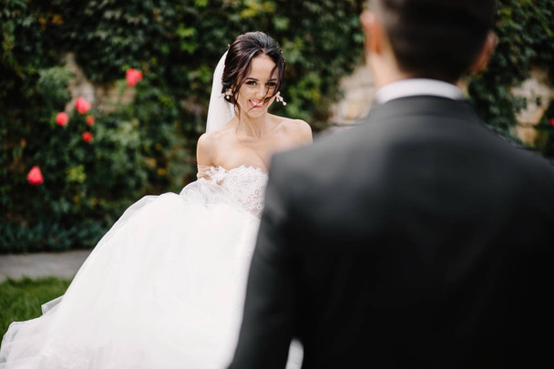 glücklich lächelnd stilvolle Braut und Bräutigam beim Spazierengehen im sommergrünen Park mit Blumenstrauß, Tanzen und Spaß am Hochzeitstag - Foto, Bild