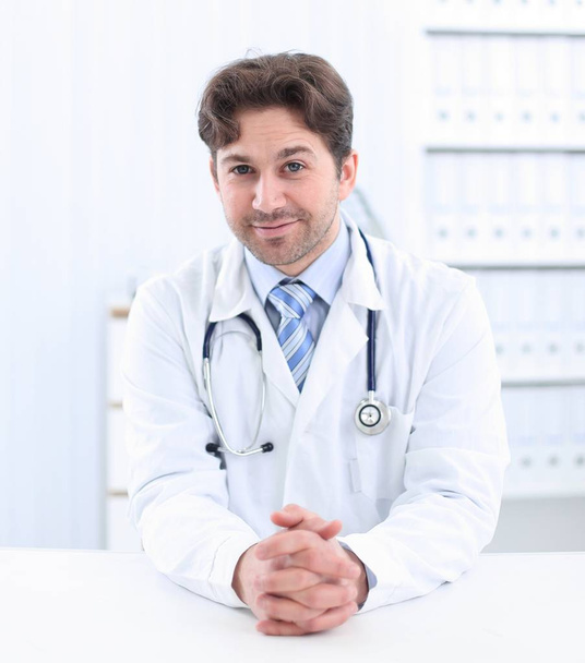 Handsome young doctor in white coat is looking at camera and smiling while standing in office - Foto, afbeelding