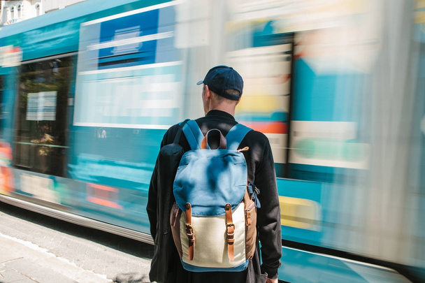 Мужчина, стоящий перед дорогой с трамваем. Мужчина турист в повседневной одежде с рюкзаком стоит перед дорогой с трамваем проходит в солнечный летний день. Move blur
. - Фото, изображение