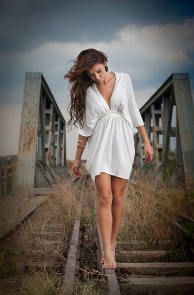 attractive woman with short white dress and long hair standing on the rails with bridge in background. Fashion  sexy girl with sexy body and long legs on the bridge posing in  white dress  - Фото, изображение