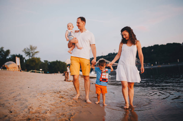 concept family beach vacation. Family of four people mom, dad and brothers children boys walking, want barefoot on the river bank by the hand at sunset in the summer - Photo, Image