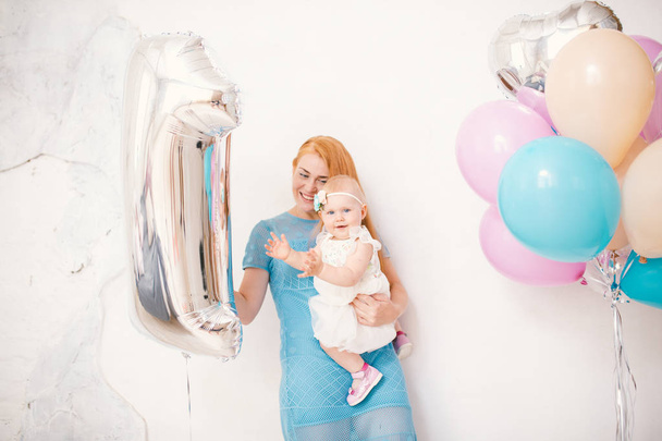 young red-haired mother holding a baby girl one year in a dress on a white background at home. The concept of a children's holiday is decorated with inflatable airy multi-colored balls - Φωτογραφία, εικόνα