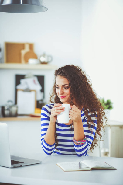 Smiling young woman with coffee cup and laptop in the kitchen at home. Smiling young woman - Foto, immagini