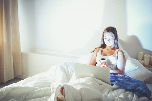 Μια όμορφη νεαρή γυναίκα με ένα laptop στο κρεβάτι και απολαμβάνοντας ένα φλιτζάνι τσάι - Φωτογραφία, εικόνα