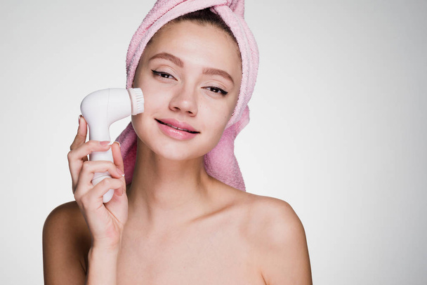χαριτωμένο νεαρή κοπέλα με μια ροζ πετσέτα στο κεφάλι της, να κάνει ένα βαθύ καθαρισμό του δέρματος του προσώπου με μια ηλεκτρική βούρτσα - Φωτογραφία, εικόνα