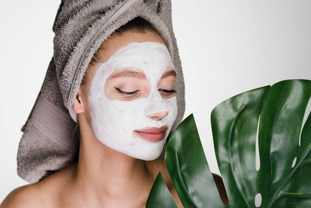 a cute young girl with a towel on her head applied a white nutritious mask to her face, holds a green leaf, day spa - Фото, зображення