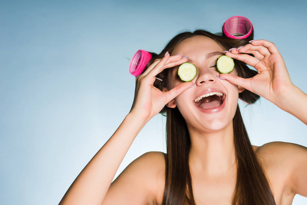 happy laughing girl holding a cucumber for moisturizing the skin of the face, on the head of curlers - Foto, afbeelding