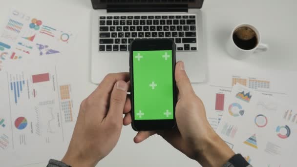 Liikemies, joka käyttää älypuhelinta, jossa on vihreä näyttö valkoisen toimiston pöydän taustalla. Huippunäkymä. Mies kädet vieritys sivuja, zoomaus, napauttamalla kosketusnäyttö. Toimiston tausta
 - Materiaali, video