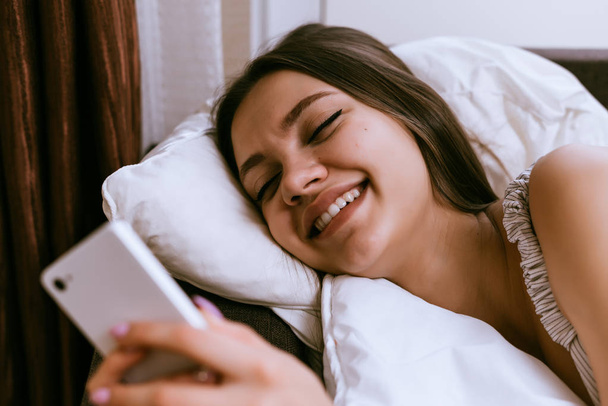 happy young girl lies in bed, looking into smartphone, smiling - Foto, imagen