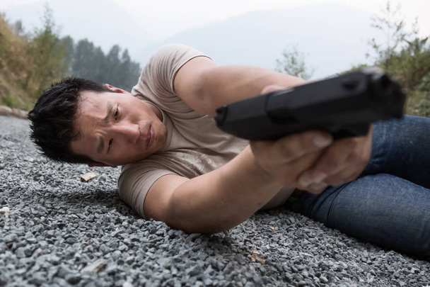 Asian young man is on the gravel ground in laying position holding a pistol and aiming to shoot at a gun range. - Foto, imagen