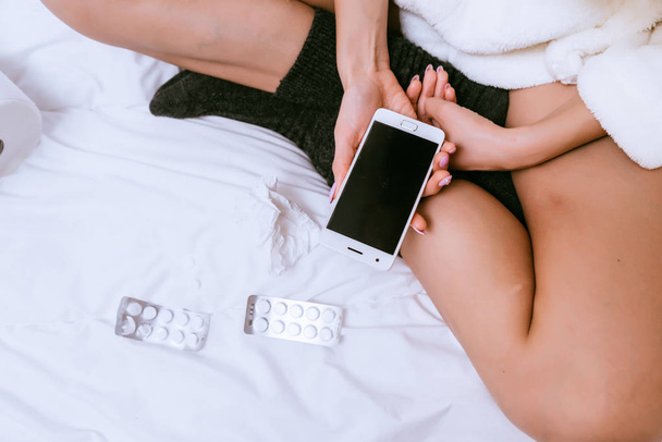 a girl in a white robe and warm socks is sitting on the bed, holding a smartphone, on the bed a lot of tablets - Photo, image
