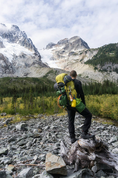 An adventurous young man wearing a big heavy backpack is standing on a log and enjoying the beautiful scenery surrounded by mountains. Taken far remote East of Vancouver and Seattle, Washington, USA. - Photo, Image