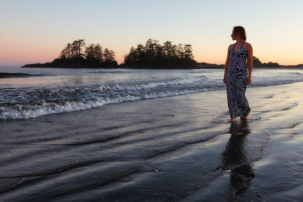 Woman wearing a casual long dress is walking on a beautiful sandy beach on Pacific Ocean Coast during a vibrant summer sunset. Taken in Tofino, Vancouver Island, British Columbia, Canada. - Φωτογραφία, εικόνα