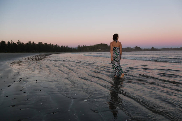 Woman wearing a casual long dress is walking on a beautiful sandy beach on Pacific Ocean Coast during a vibrant summer sunset. Taken in Tofino, Vancouver Island, British Columbia, Canada. - Foto, Bild