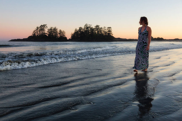 Kobieta nosi strój długi jest chodzenie na pięknej, piaszczystej plaży na wybrzeżu Pacyfiku podczas Letnich wibrujący zachód słońca. Podjęte w Tofino, Wyspa Vancouver, British Columbia, Kanada. - Zdjęcie, obraz