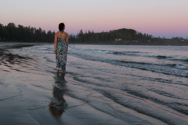 Woman wearing a casual long dress is walking on a beautiful sandy beach on Pacific Ocean Coast during a vibrant summer sunset. Taken in Tofino, Vancouver Island, British Columbia, Canada. - Valokuva, kuva