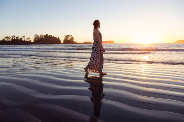 Woman wearing a casual long dress is walking on a beautiful sandy beach on Pacific Ocean Coast during a vibrant summer sunset. Taken in Tofino, Vancouver Island, British Columbia, Canada. - Zdjęcie, obraz