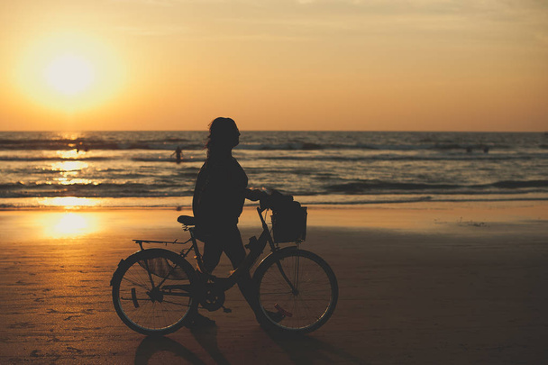 A woman makes an evening bike ride along the sea. People walk along the sea at sunset. Sunset on sea. Silhouette of people. Travelers at sunset. Tourists on vacation - Photo, image