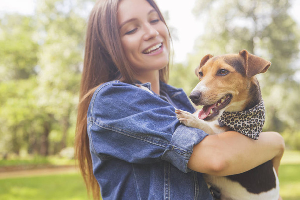 Χαριτωμένο όμορφη slim Καυκάσιος μελαχρινή γυναίκα με απλό ντύσιμο (φούστα, πουκάμισο και τζιν σακάκι) μια βόλτα στο πάρκο στην θερινή ημέρα με το αξιολάτρευτο Τζακ Ράσελ σκυλί. Αυτοί γεμάτη χαρά, χαμογελώντας και να διασκεδάσουν - Φωτογραφία, εικόνα