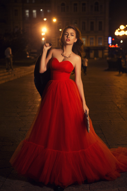 Beautiful sexy caucasian woman with long curly brunette hair and make up on big eyes and full sensual lips. She coming back from party, walking in evening dress on european street with traffic lights - Foto, Bild
