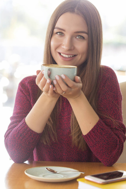 Morena joven mujer caucásica en suéter casual y jeans sentados en la cafetería, sonriendo felizmente con una taza de café. Retrato de estilo de vida en interiores. Copiar espacio
 - Foto, Imagen