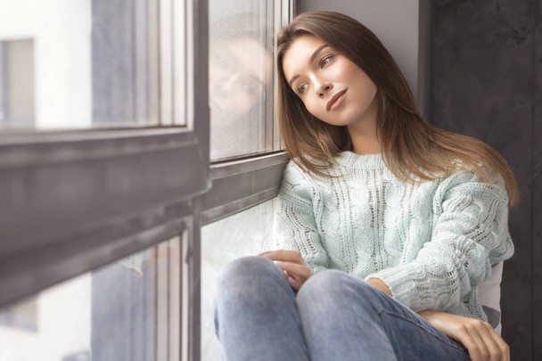 Cute adult young woman sitting near window in casual clothes (sweater and jeans), looking sideways thoughtful. Copy space - Foto, imagen