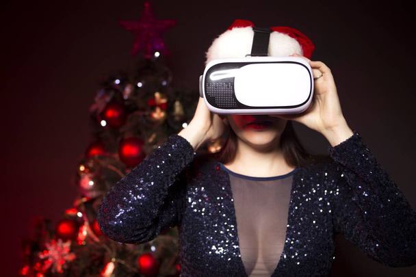 Cute smiling woman in Christmas hat and evening sexy dress using VR glasses. New Year tree with red light on a background - Photo, Image