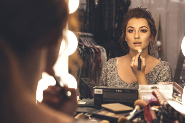 Brunette woman applying make up (paint her skin) for a evening date in front of a mirror. Focus on her reflection - 写真・画像