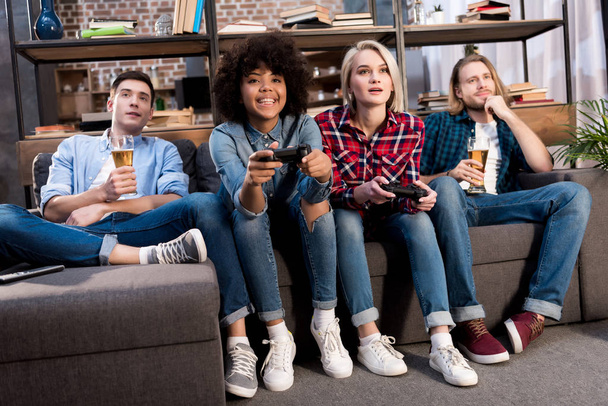 multikulturní dívky hrát videohry na pohovce doma, muži popíjeli pivo - Fotografie, Obrázek